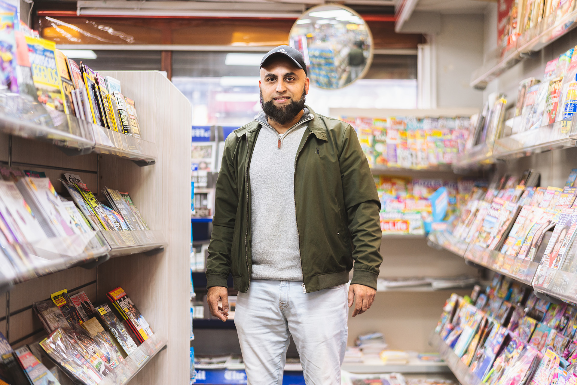 Photo portrait of a man standing inside a newsagents. On either side are rows of magazines. The man wears a cap and jacket over his clothes, and smiles at the camera.