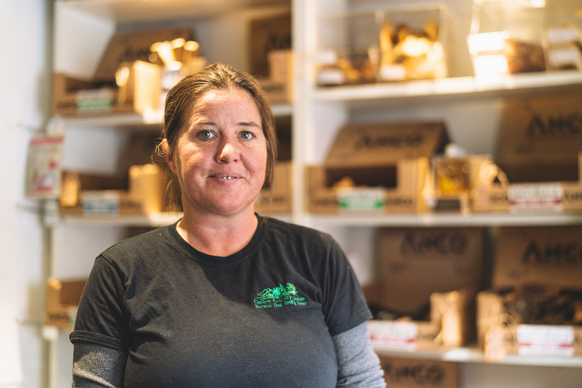 Photograph of a woman with brown hair inside a shop or workspace. In the background are shelves filled with packaging.