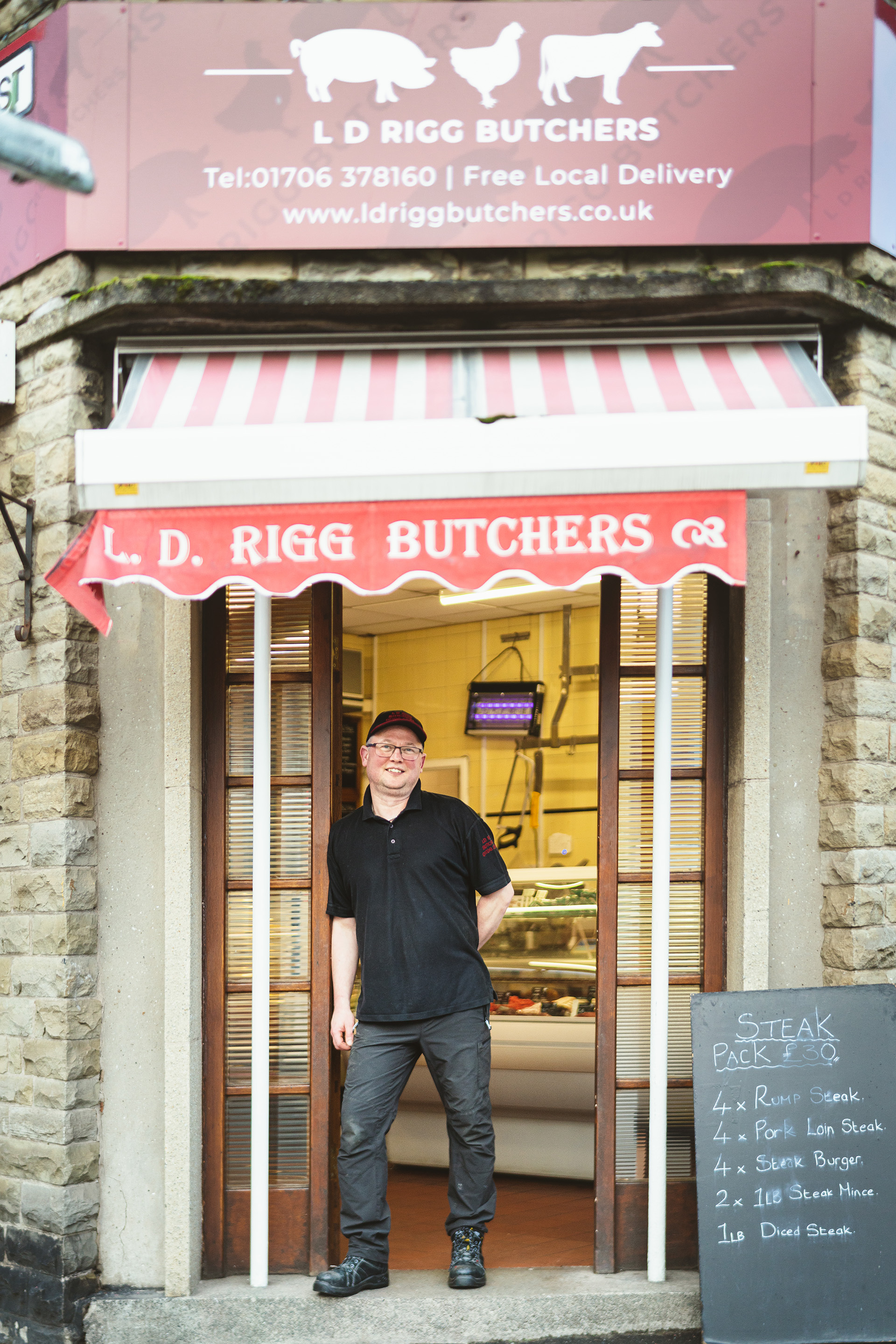 Photograph of a man standing in the doorway of L. D. Rigg Butchers. He leans on the doorway and smiles towards the camera.