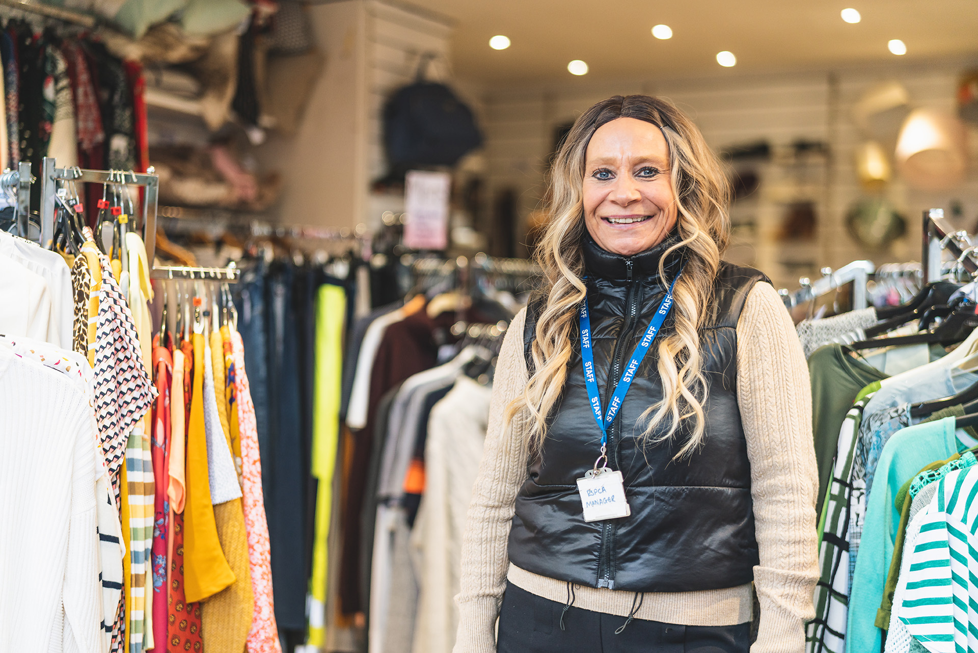 Photograph of a blonde woman standing next to rails of colourful clothing. She smiles at the camera.