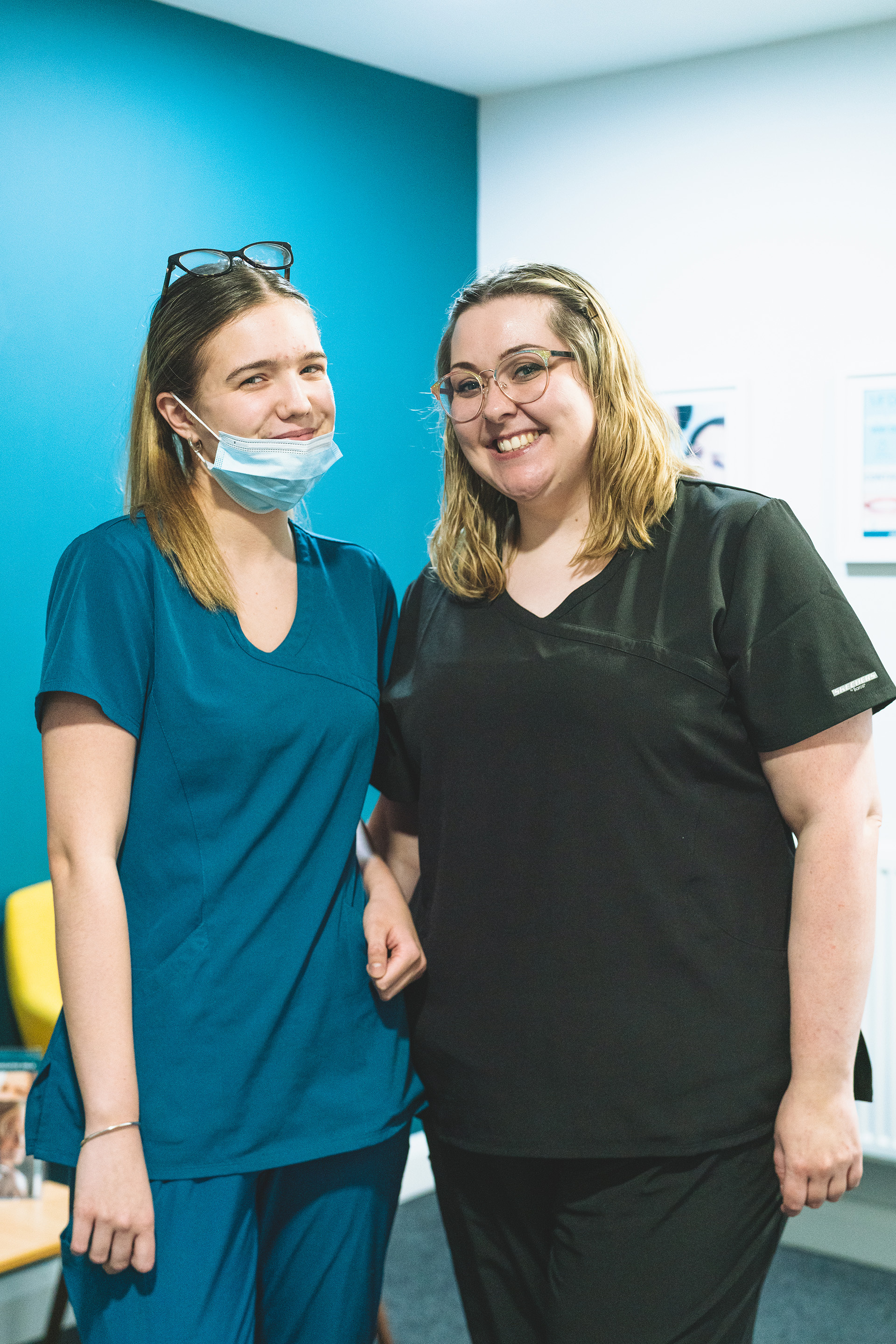 Two blonde women stand side by side inside a clinic. One wears a blue face mask.