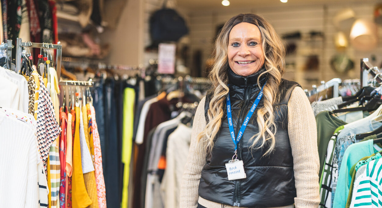 Photograph of a blonde woman standing next to rails of colourful clothing. She smiles at the camera.